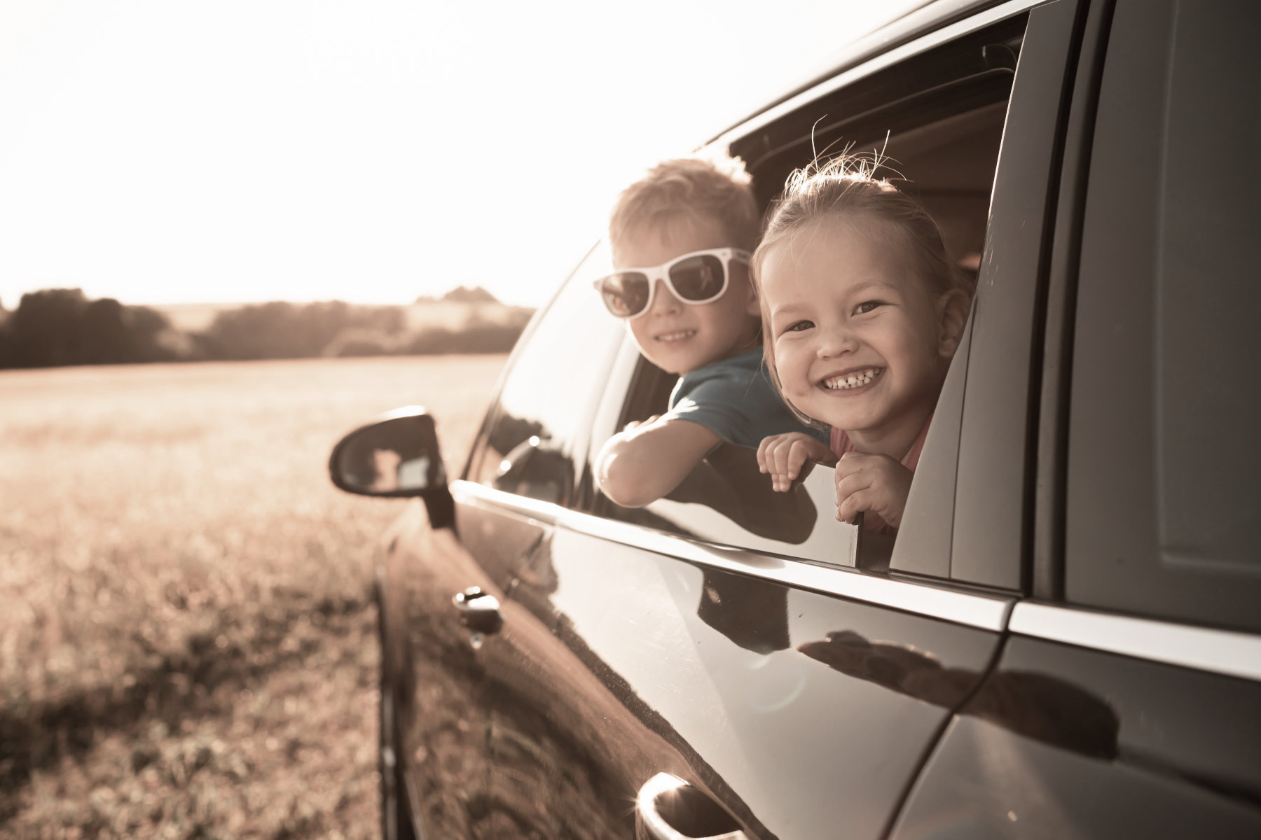 Two children leaning out the window from the back seat of a car.