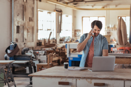 Commercial Property Insurance in MN - A man in a workshop talking on a cell phone and using a laptop computer.